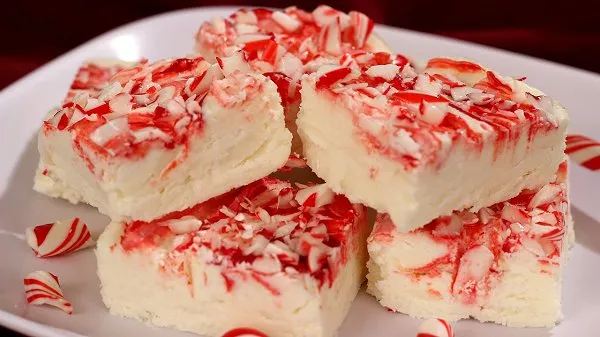 Creamy, peppermint flavored white fudge with crushed candy canes on top stacked on a white plate.