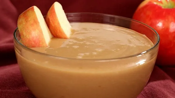 Creamy caramel apple dip in a clear bowl with apple slices dipped halfway, on a white plate with an apple in the background.