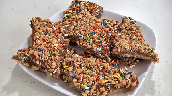 Chocolate covered saltine crackers topped toffee pieces, chopped nuts and colorful sprinkles stacked on a white plate.