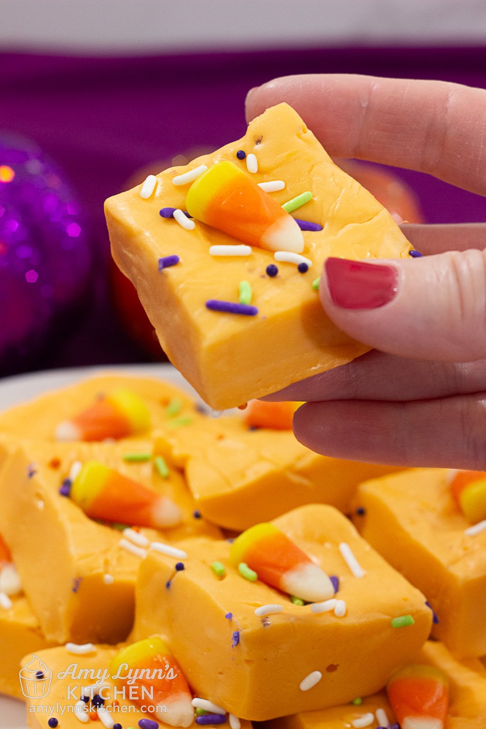 Hand holding a square of Easy Halloween Orange Fudge topped with candy corn and colorful sprinkles, with more fudge pieces in the background.