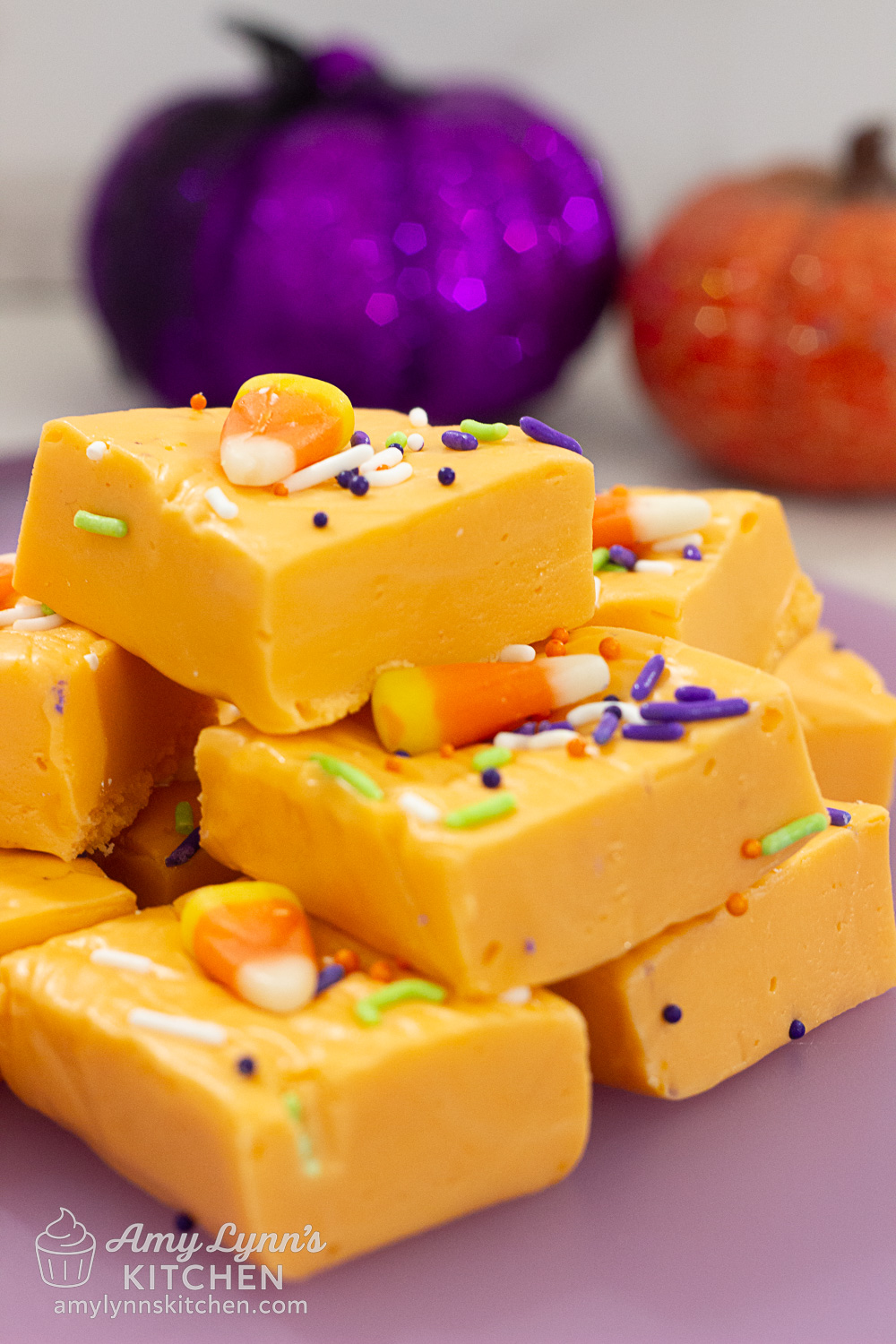 Easy Halloween Orange Fudge squares topped with candy corn and colorful sprinkles, with sparkly purple and orange pumpkins in the background.