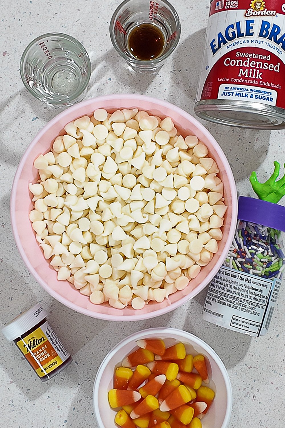 Ingredients for Easy Halloween Orange Fudge including white chocolate chips, sweetened condensed milk, orange gel food coloring, candy corn, sprinkles, and extracts on a kitchen counter.