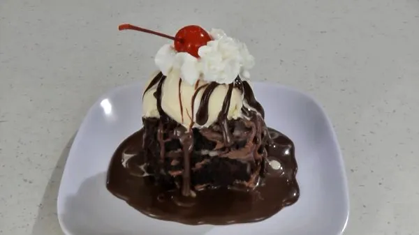 Slice of chocolate cake topped with vanilla ice cream, whipped cream, a cherry, and drizzled with hot fudge sauce.