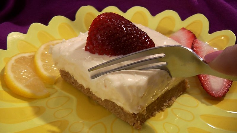 Wide shot of creamy no-bake lemon cheesecake squares on a graham cracker crust, with whipped cream and strawberry garnish