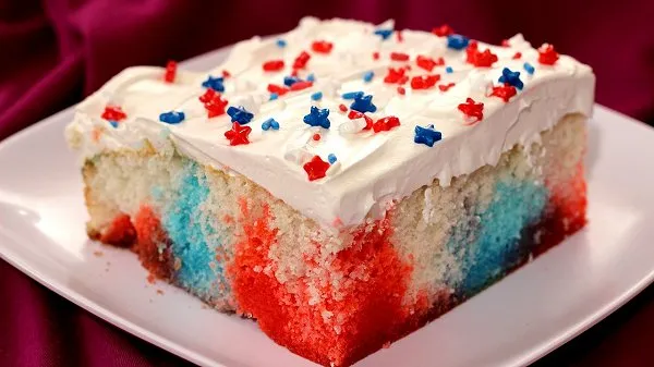 Slice of Easy Red, White and Blue Jello Poke Cake on a white plate, with vibrant red and blue Jello streaks inside fluffy white cake, topped with whipped cream and festive sprinkles
