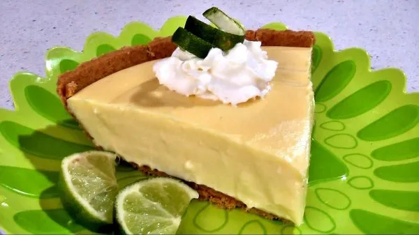 A slice of key lime pie topped with whipped cream and lime zest on a green plate surrounded by slices of lime.