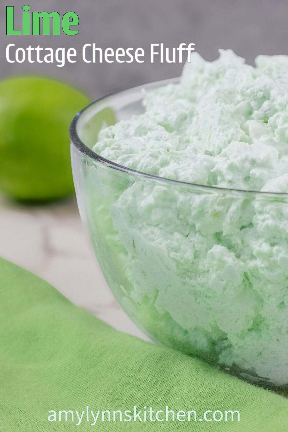 A bright and fluffy cottage cheese Jell-O side dish in a clear glass bowl, surrounded by a fresh lime and a green cloth on a light colored background. A light and creamy retro dessert made with lime Jell-O, pineapple, and whipped topping.