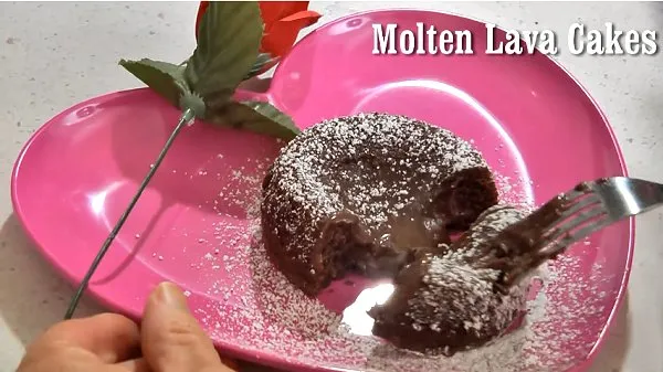Molten lava cake on a pink heart-shaped plate, cut open to show the gooey chocolate center, dusted with powdered sugar and topped with a red rose.