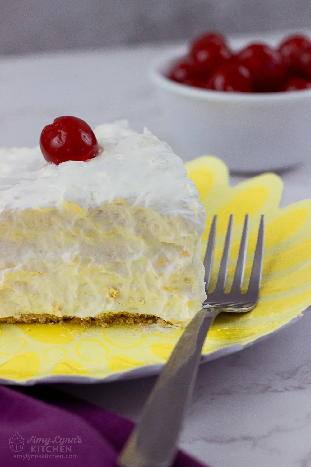 Close-up of a creamy slice of No Bake Pineapple Cream Cheese Pie with a graham cracker crust and maraschino cherry on top, served on a yellow plate with a fork.