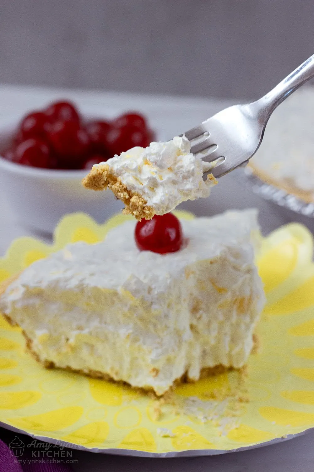 Fork holding a bite of creamy No Bake Pineapple Cream Cheese Pie with graham cracker crust, above a slice topped with a maraschino cherry on a yellow plate.