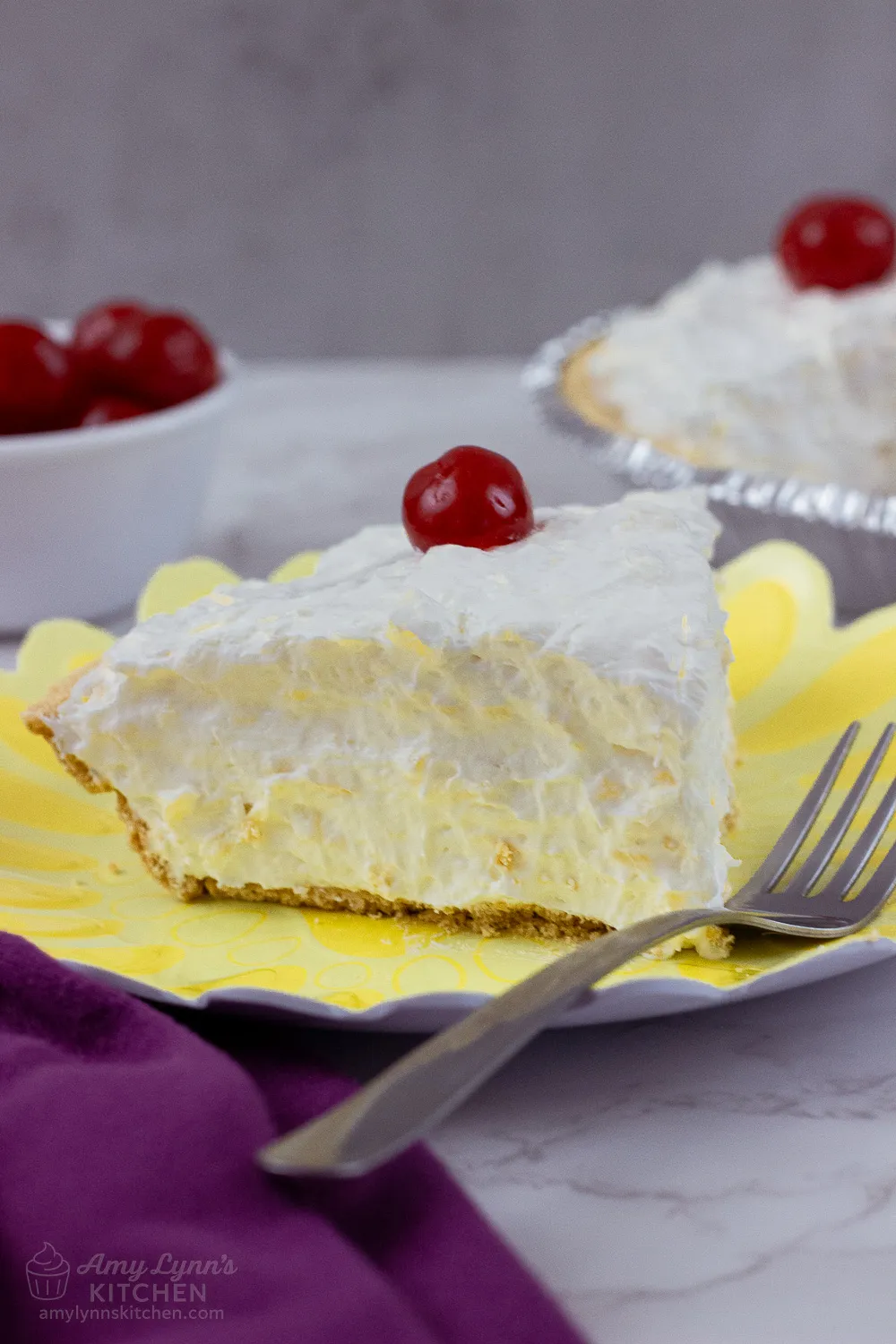 A close-up slice of creamy No Bake Pineapple Cream Cheese Pie on a yellow plate, topped with a bright red maraschino cherry, with a graham cracker crust and fluffy pineapple filling, served with a fork.