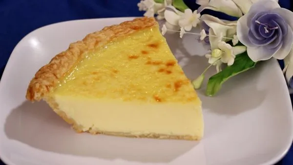 A big slice of old fashioned custard pie on a white plate with flowers in the background.