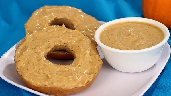 Bagel halves spread with pumpkin cream cheese on a white plate, with a small bowl of extra pumpkin cream cheese and a pumpkin in the background.