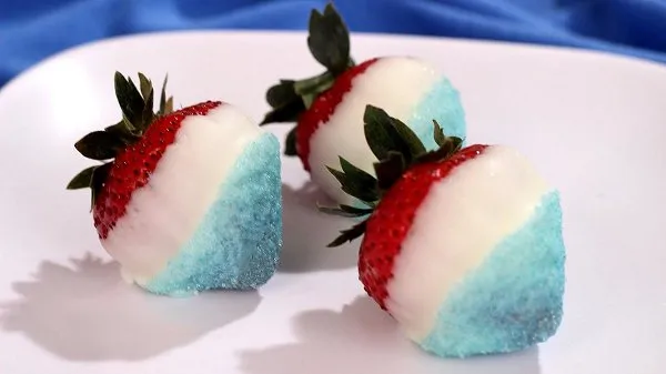 Red, white, and blue strawberries coated with white chocolate and blue sugar on a white plate.