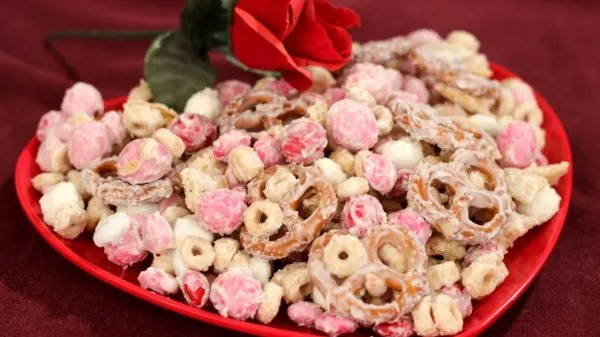A red heart plate nearly overflowing with Valentine's Day party mix and a single red rose in the background.